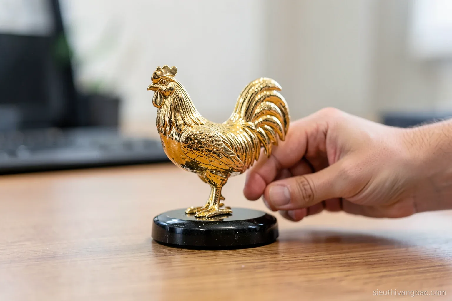 Side view of a gold-plated rooster figurine mounted on a polished black stone base, a hand gently testing it on a wooden desk, soft diffused office light