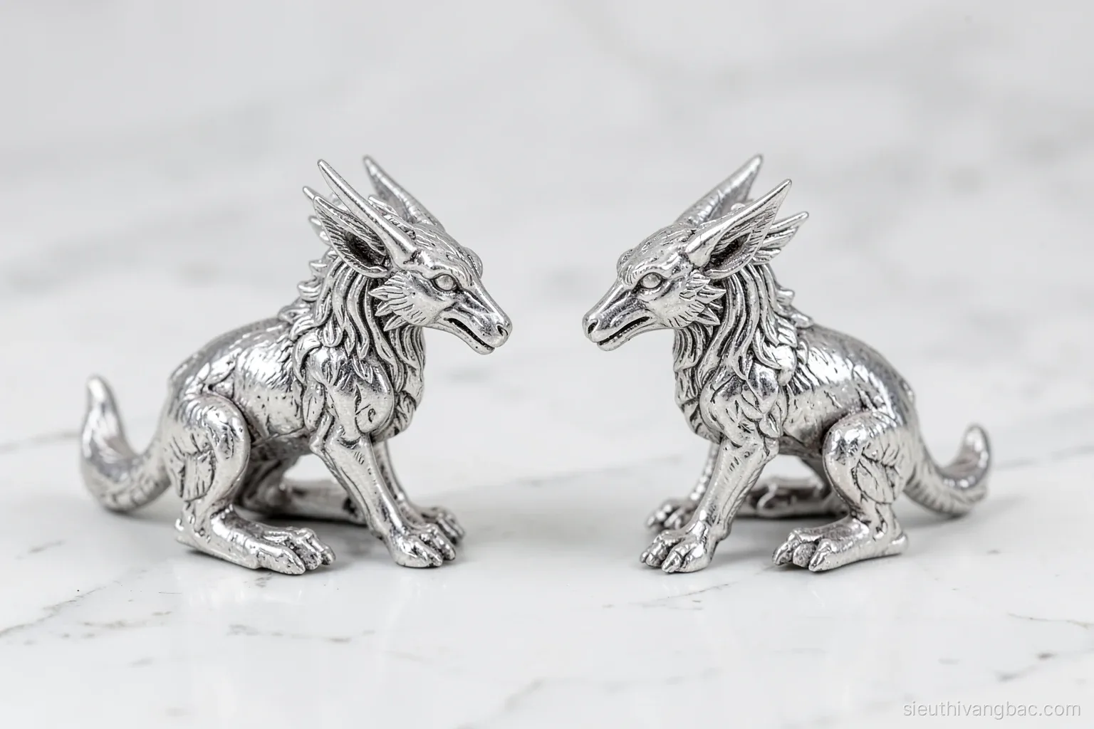 Two matching silver-colored mythical creature figurines facing each other on a white marble tabletop, symmetrical composition, eye-level shot, soft even studio daylight