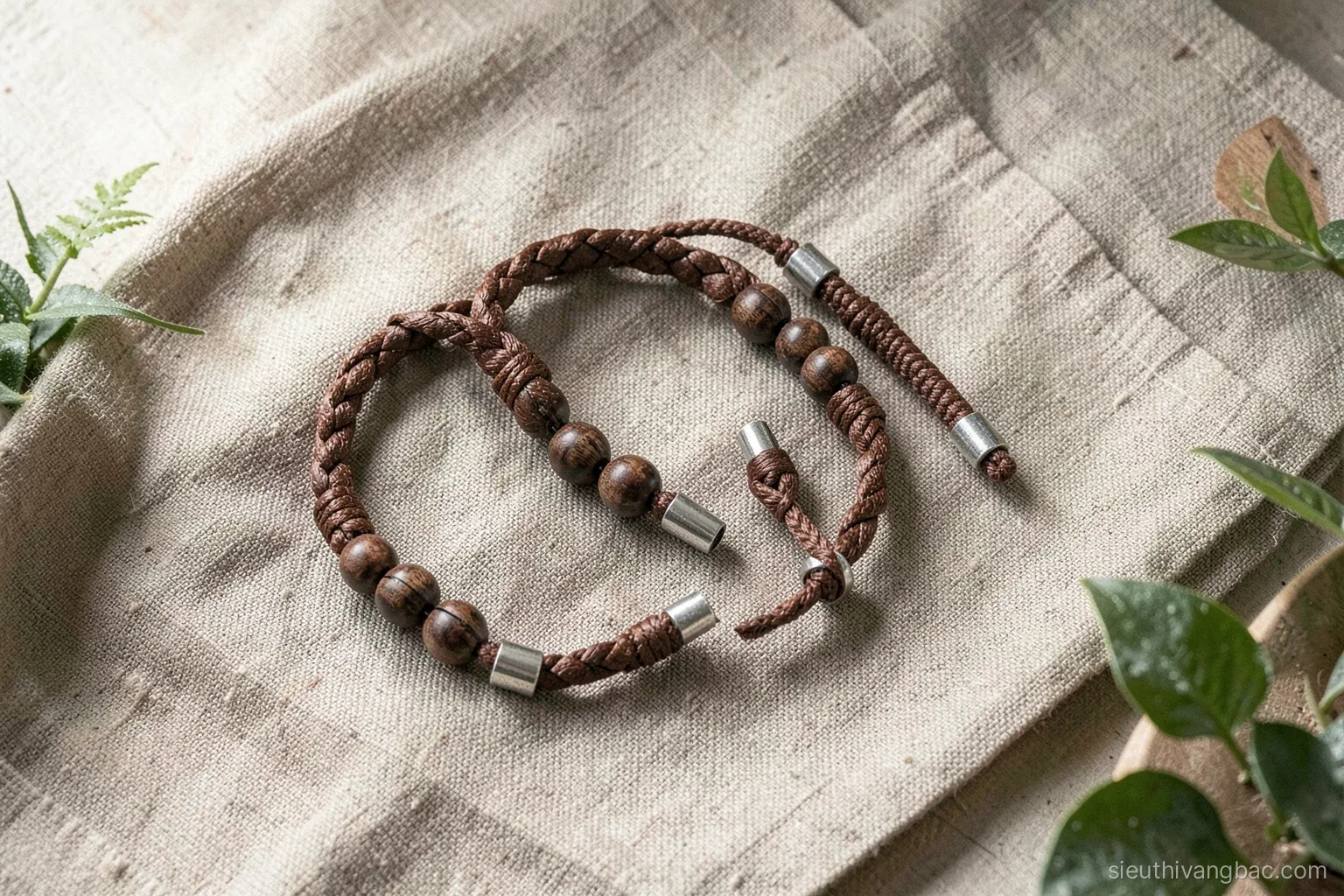 Flat lay of men's bracelets combining braided brown leather, dark wooden beads and paracord with silver end caps on rough linen, overhead natural light, earthy palette